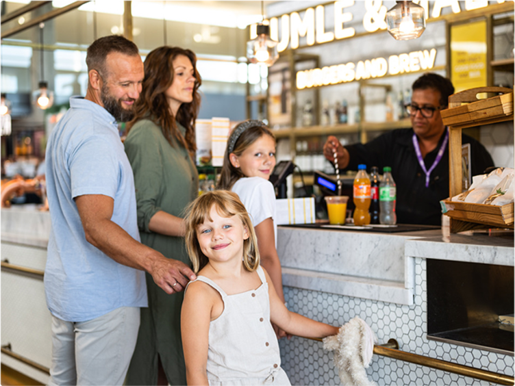 A family of four orders food in a modern café, with smiling children and parents, while an employee serves beverages in a bright, friendly environment.