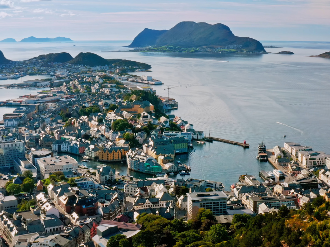 Aerial view of a coastal city with a harbor, colorful buildings, and surrounding islands and mountains on a calm fjord-like bay