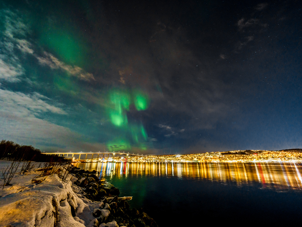 Northern lights over a snowy waterfront city at night with a lit bridge and reflections on calm water