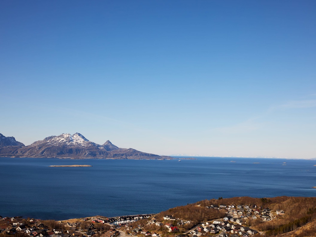 Panoramic coastal landscape with blue ocean, snow-capped mountains, and a small seaside village under a clear sky