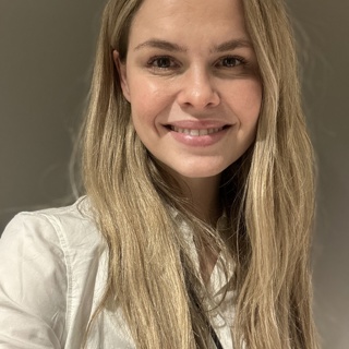 Smiling woman with long blond hair wearing a black top, seated indoors with a plant in the background
