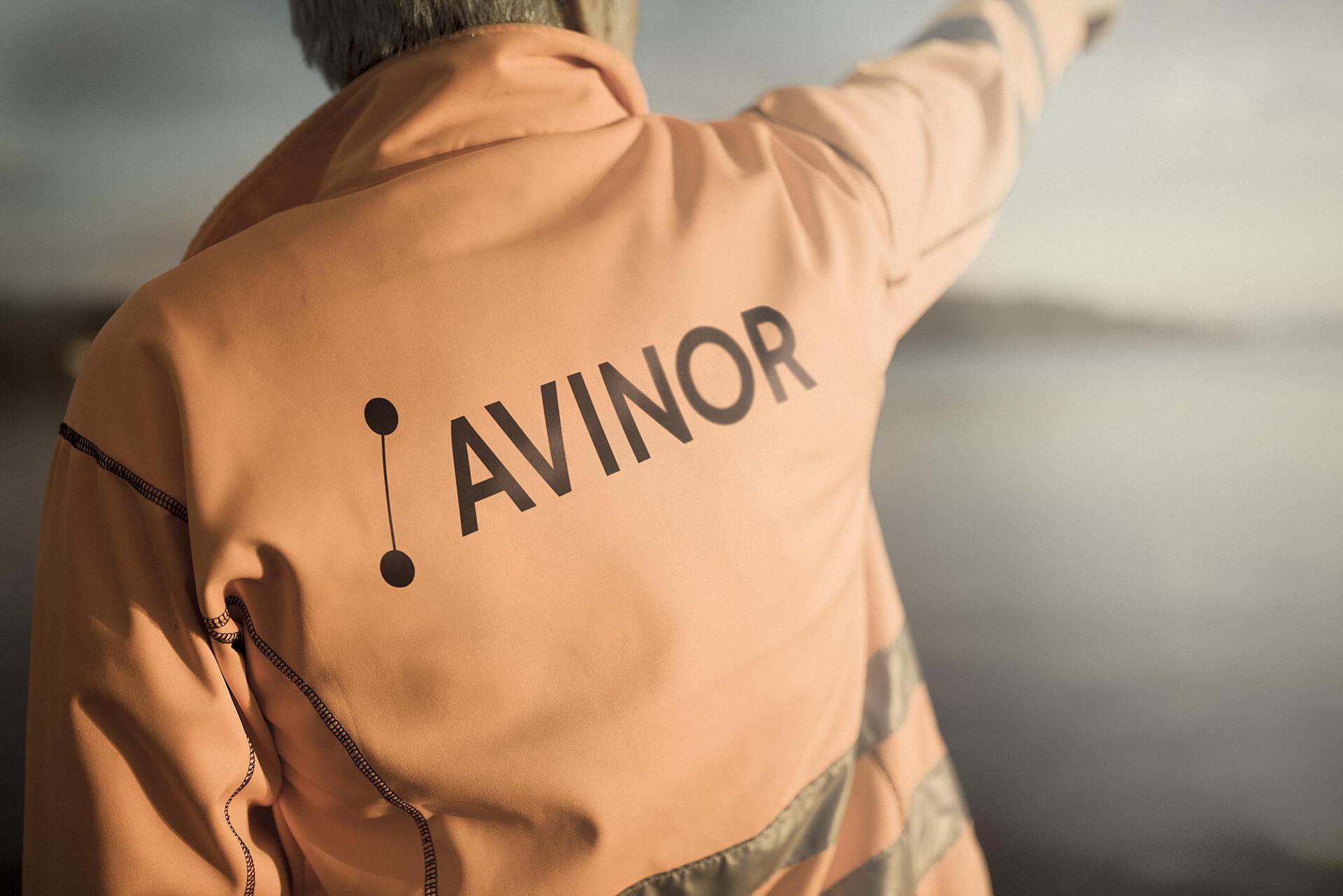 Person wearing an orange jacket with the Avinor logo, pointing towards the horizon by a waterfront.