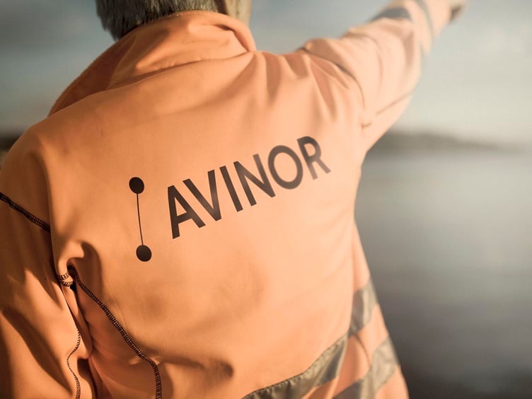Person wearing an orange jacket with the Avinor logo, pointing towards the horizon by a waterfront.