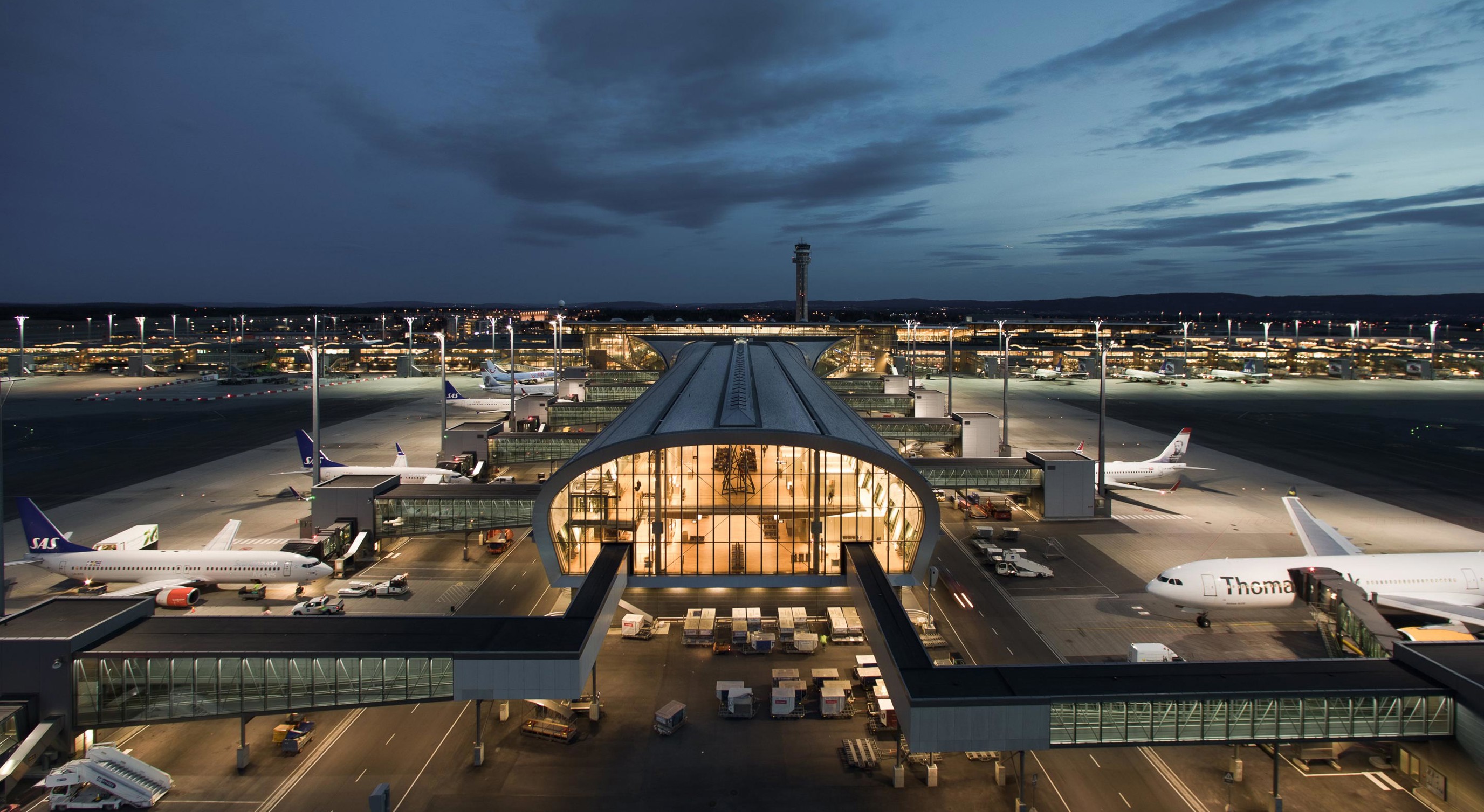 Flyplass ved skumring med opplyste terminaler og parkerte fly, omgitt av en opplyst rullebane og en mørk nattehimmel