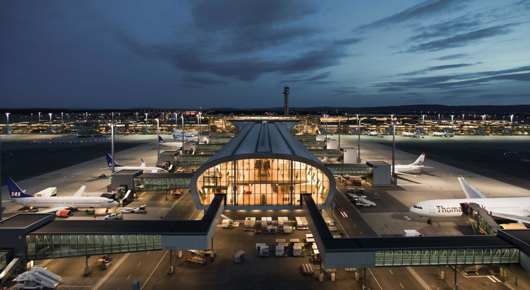 Flyplass ved skumring med opplyste terminaler og parkerte fly, omgitt av en opplyst rullebane og en mørk nattehimmel