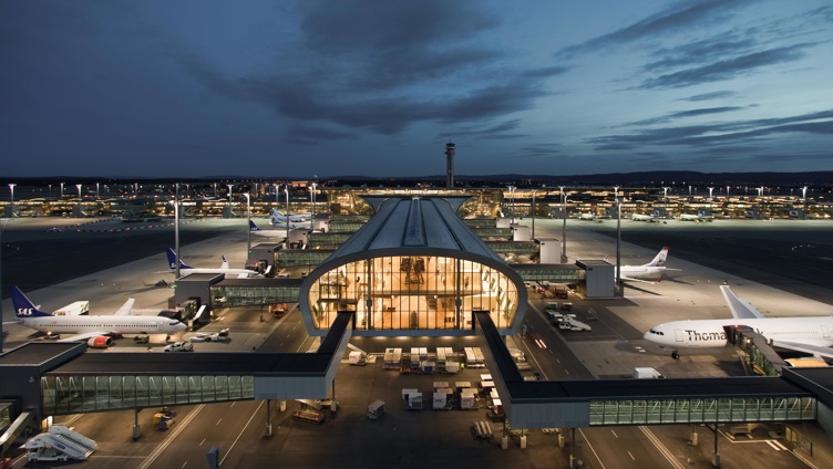 Flyplass ved skumring med opplyste terminaler og parkerte fly, omgitt av en opplyst rullebane og en mørk nattehimmel