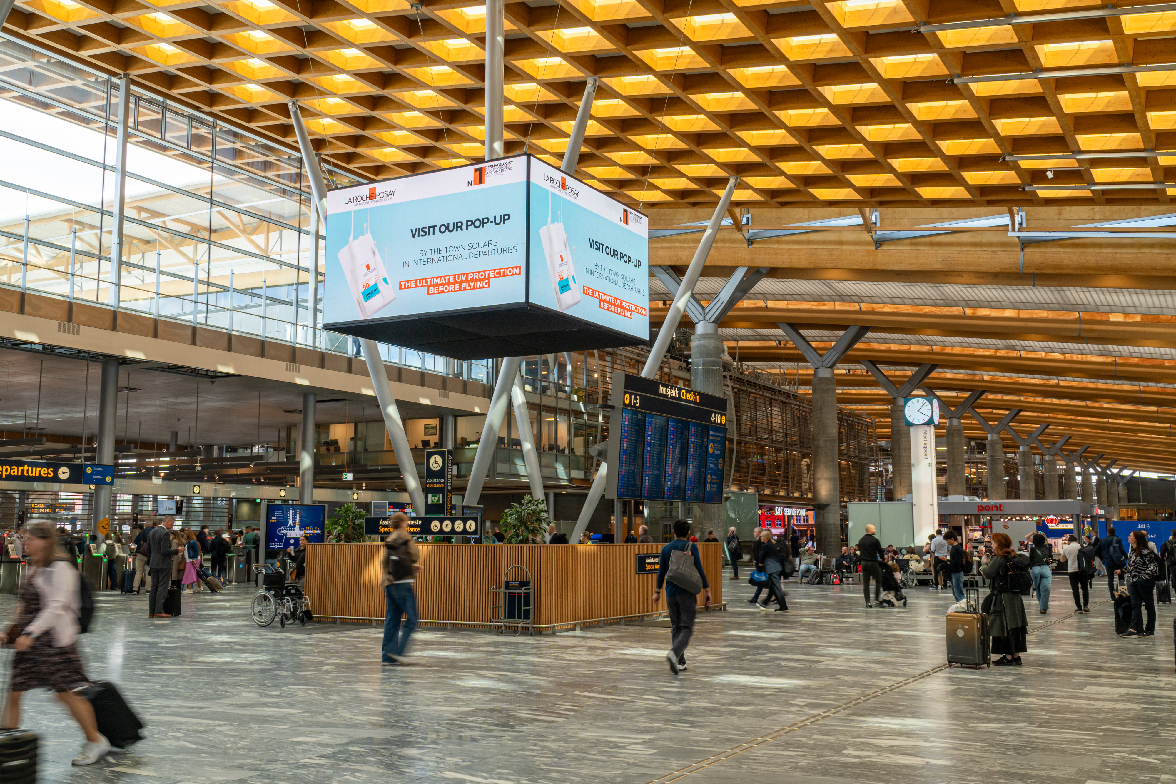 Flyplassterminal med reisende, informasjonsskjermer og reklameplakat for popup-butikk. Moderne arkitektur med store vinduer og trepanel i taket