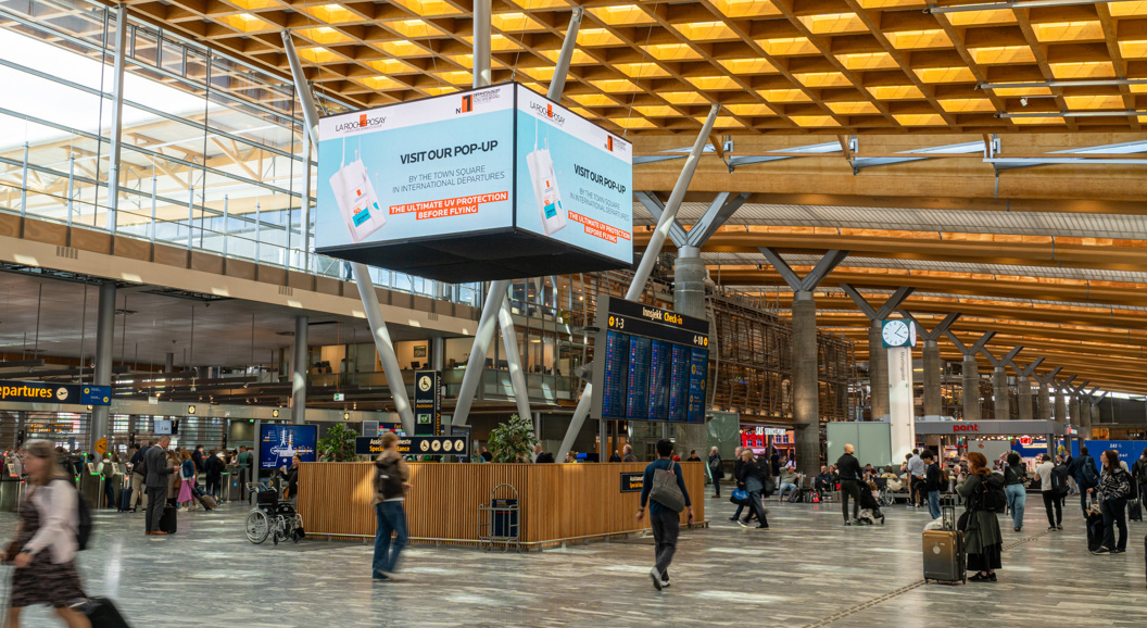 Flyplassterminal med reisende, informasjonsskjermer og reklameplakat for popup-butikk. Moderne arkitektur med store vinduer og trepanel i taket
