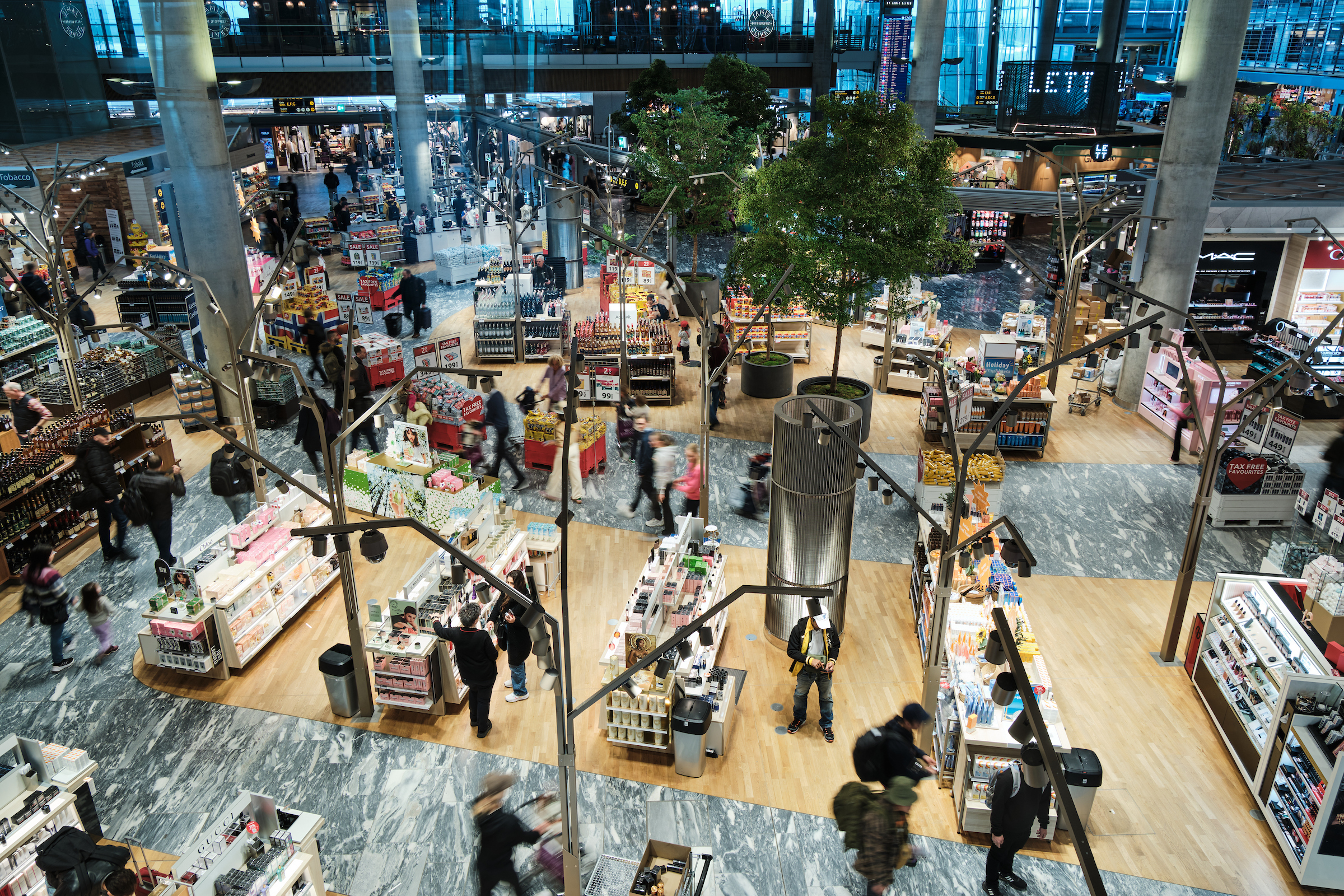 Interiør av en travel flyplassbutikk med folk som handler, omgitt av hylleutstillinger med kosmetikk, parfyme og andre varer. Butikken har moderne innredning med trær og åpen planløsning