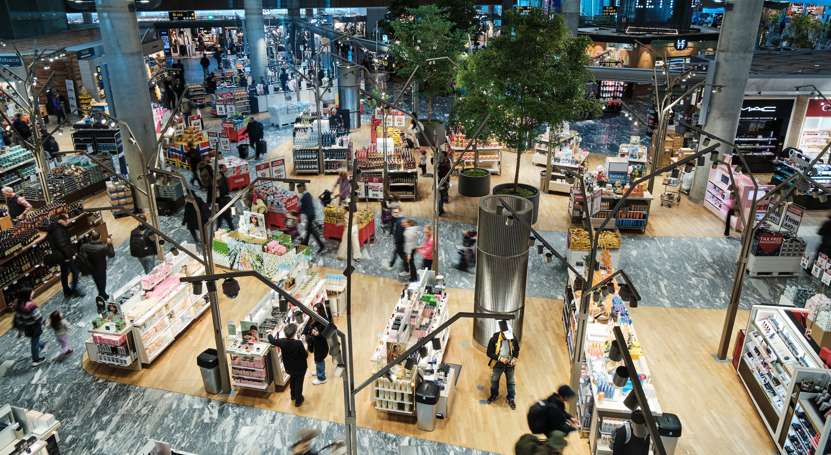 Flyplassanlegg med travle shoppingsoner, moderne innredning, og grønne planter; passasjerer handler i duty-free områder ved siden av kosmetikk- og matvareseksjoner