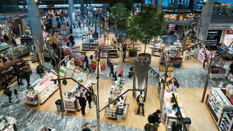 Flyplassanlegg med travle shoppingsoner, moderne innredning, og grønne planter; passasjerer handler i duty-free områder ved siden av kosmetikk- og matvareseksjoner