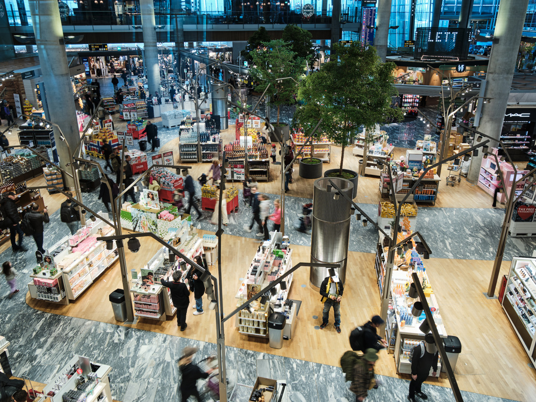 Interiør av en travel flyplassbutikk med folk som handler, omgitt av hylleutstillinger med kosmetikk, parfyme og andre varer. Butikken har moderne innredning med trær og åpen planløsning