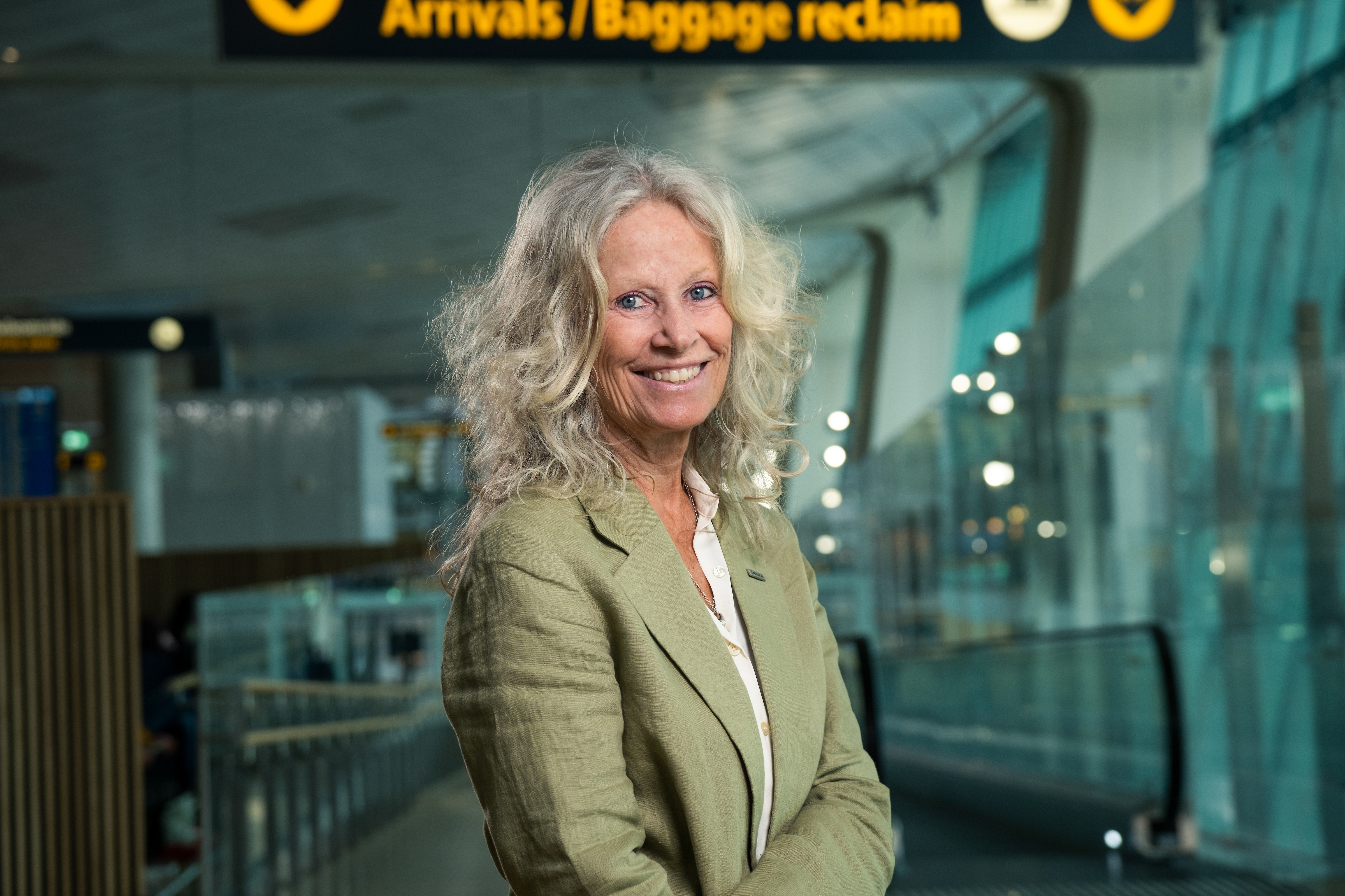 Kvinne som smiler ved ankomstområdet på flyplassen med skilt for bagasjeutlevering i bakgrunnen