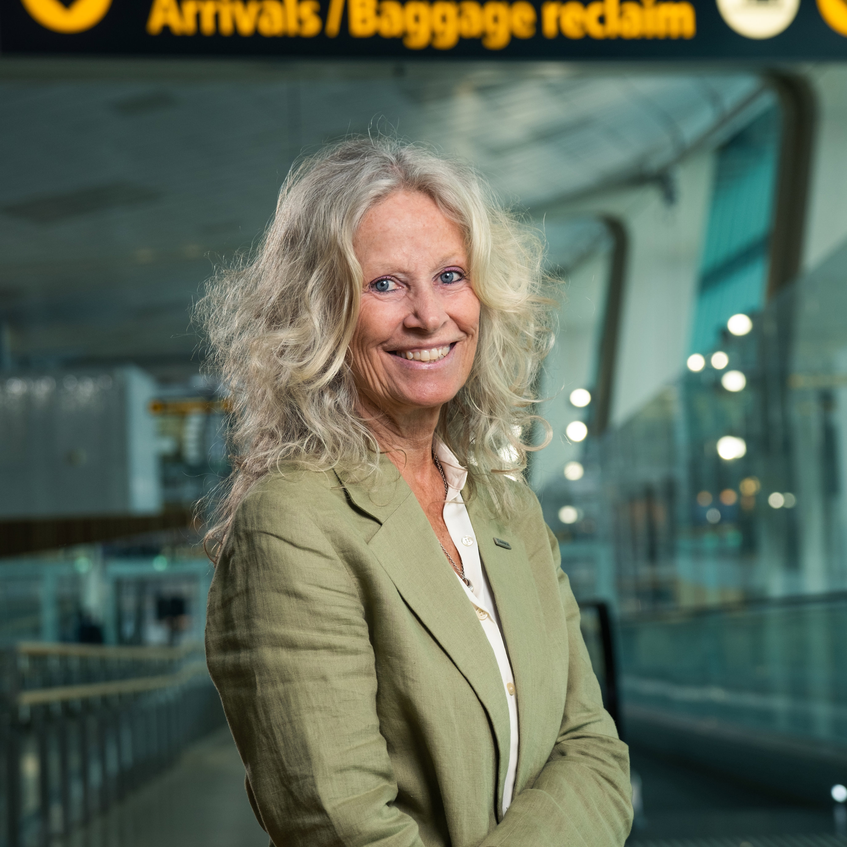 Kvinne som smiler ved ankomstområdet på flyplassen med skilt for bagasjeutlevering i bakgrunnen