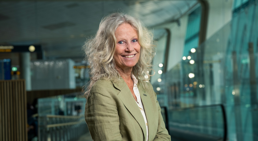 Kvinne som smiler ved ankomstområdet på flyplassen med skilt for bagasjeutlevering i bakgrunnen