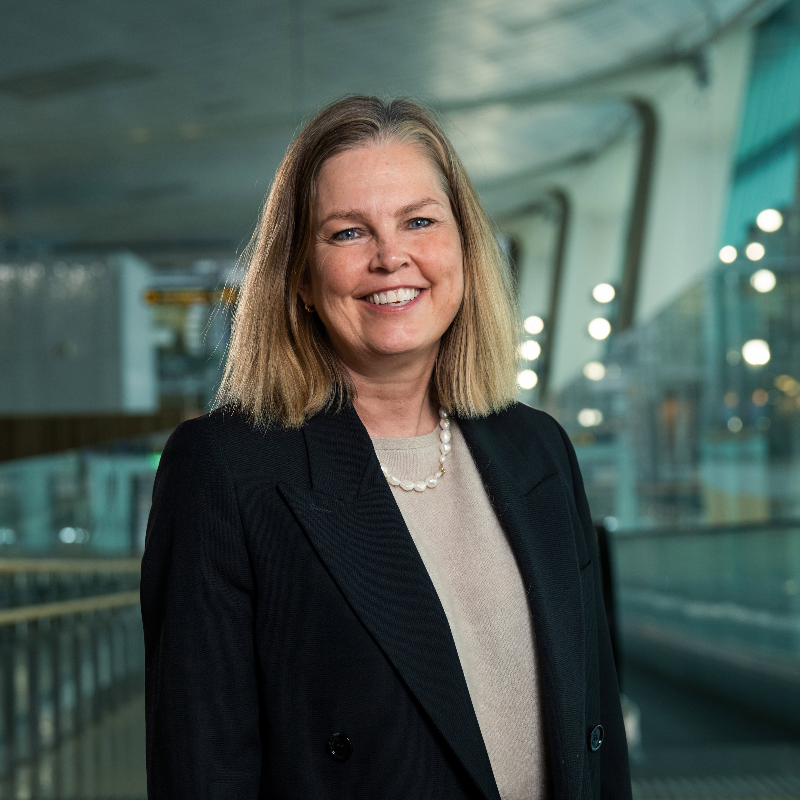 Smilende kvinne i svart blazer som står i en moderne innendørs setting med glass og lys i bakgrunnen