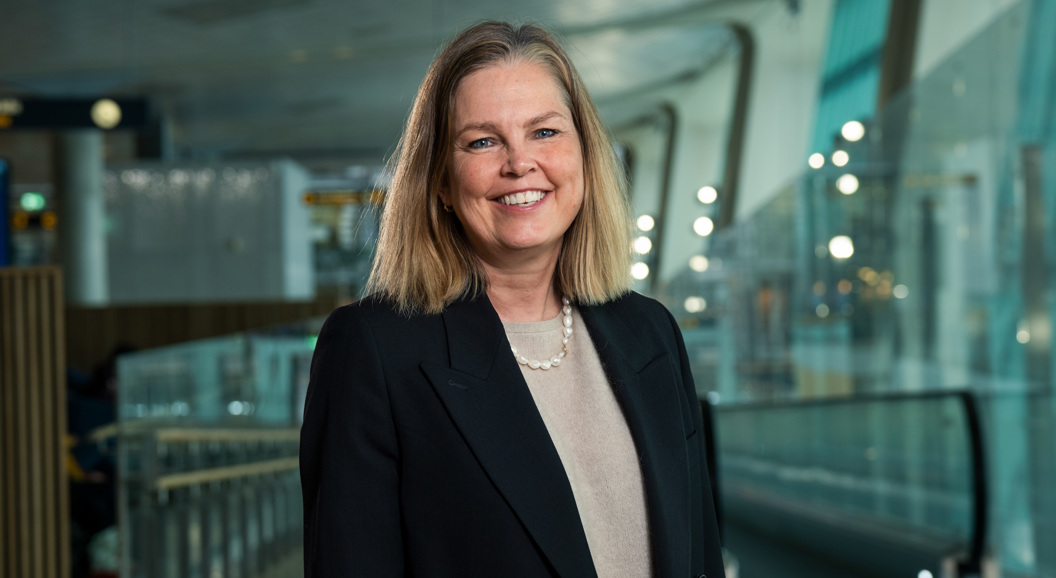 Smilende kvinne i svart blazer som står i en moderne innendørs setting med glass og lys i bakgrunnen