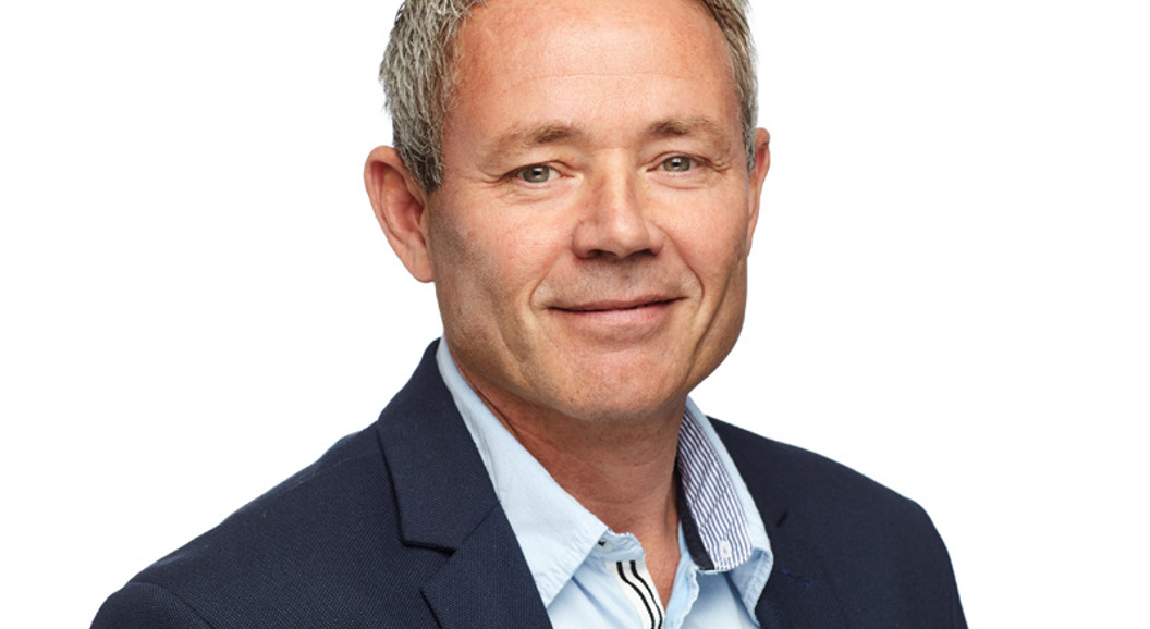 Mann i dressjakke og lyseblå skjorte smiler mot kameraet, med en nøytral bakgrunn.