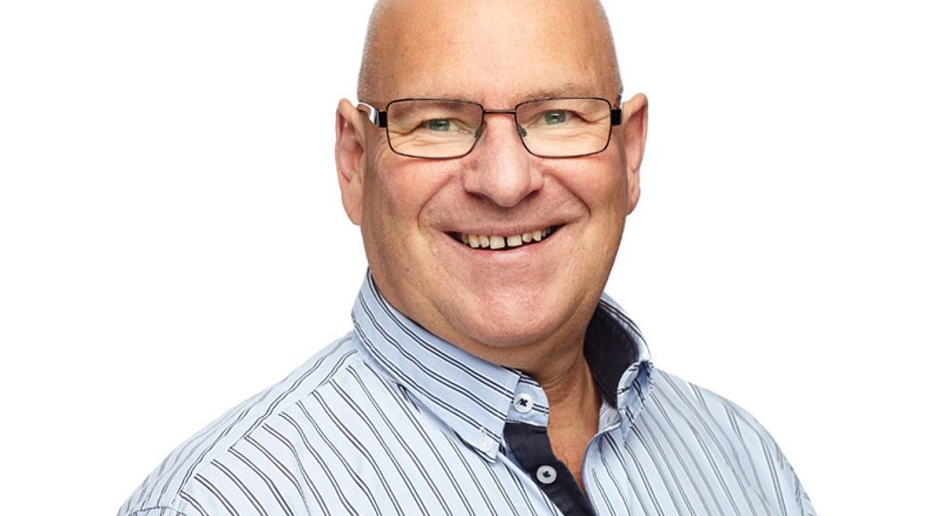 Mann med briller og stripete skjorte smiler mot kamera, hvit bakgrunn.