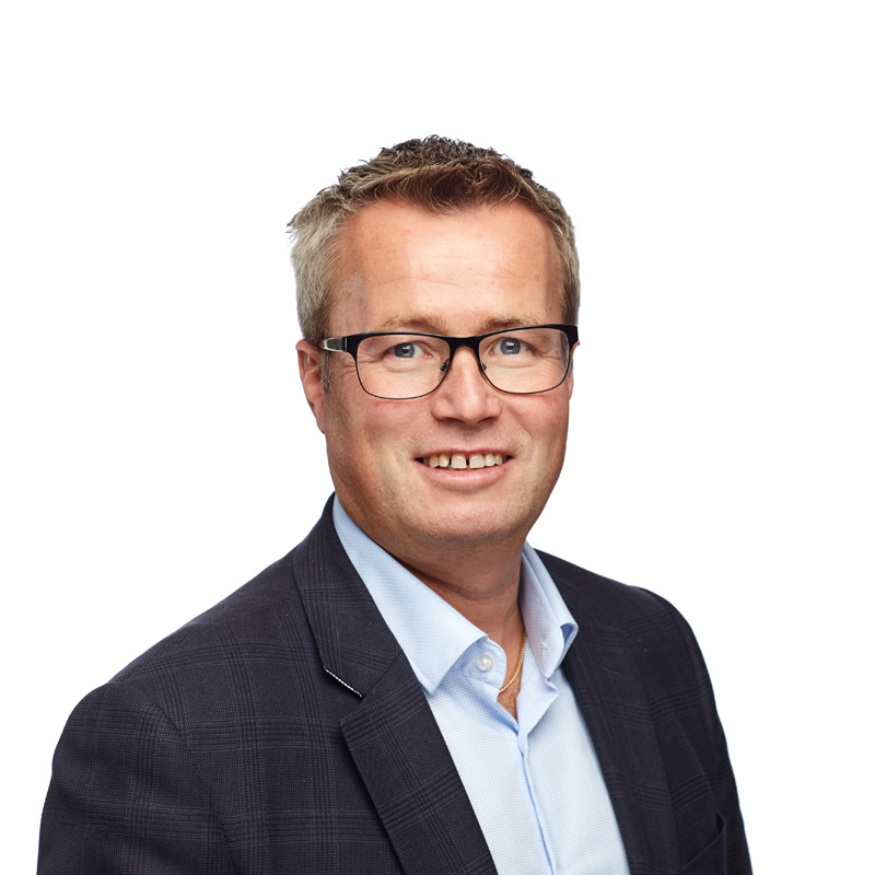 En smilende mann med briller iført dressjakke og lyseblå skjorte, med hvit bakgrunn.