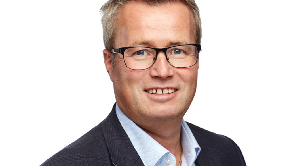 En smilende mann med briller iført dressjakke og lyseblå skjorte, med hvit bakgrunn.