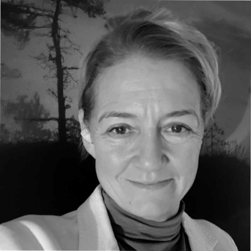 En kvinne smiler mot kameraet, iført jakke og høyhalset genser, med trær i bakgrunnen. Bildet er i svart-hvitt.