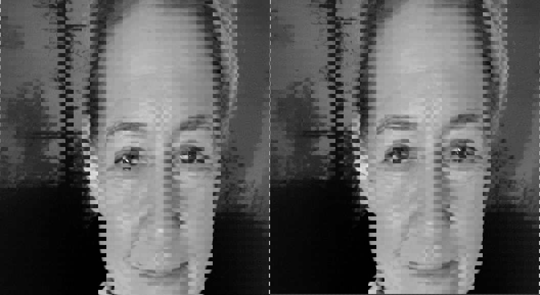 En kvinne smiler mot kameraet, iført jakke og høyhalset genser, med trær i bakgrunnen. Bildet er i svart-hvitt.