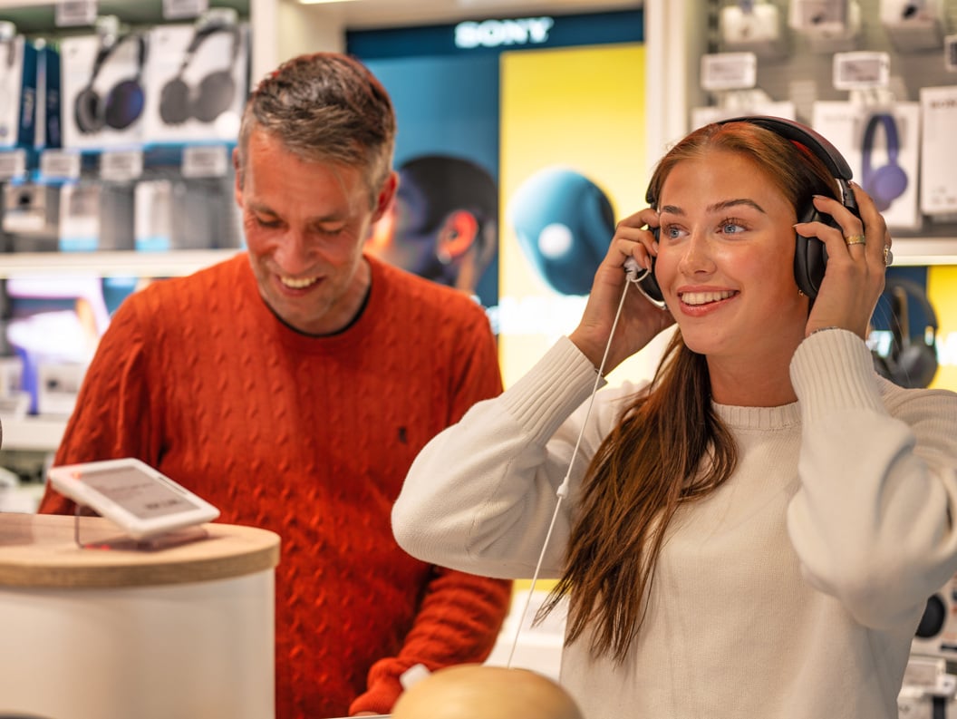 Kvinne prøver hodetelefoner i elektronikkbutikk mens en mann i rød genser smiler ved siden av
