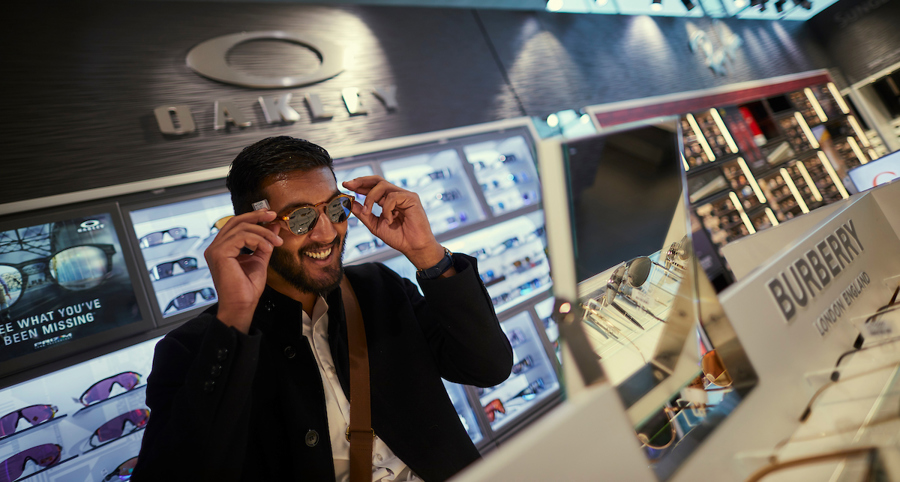 A man tries on sunglasses in an optician's store, with the Oakley logo in the background.