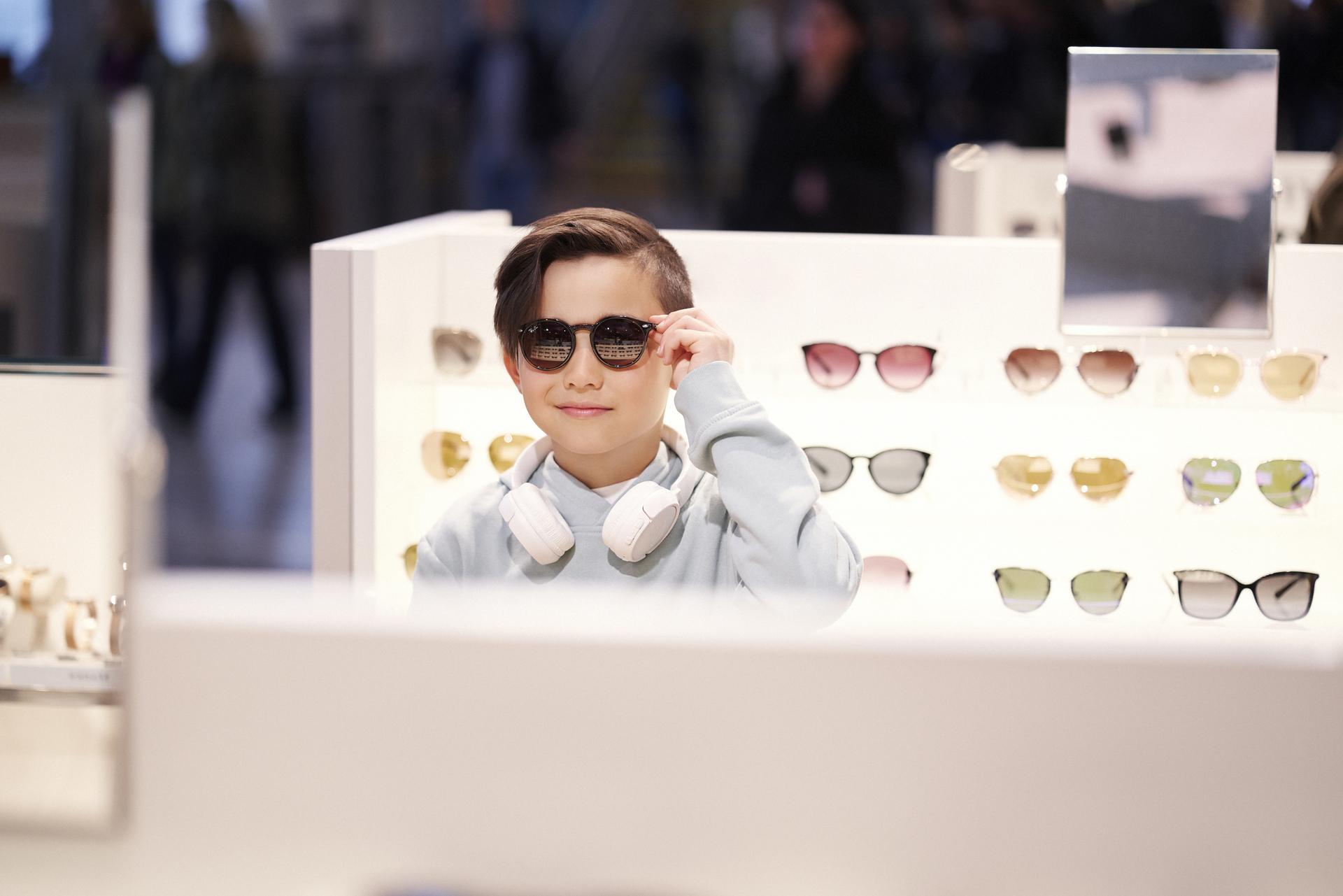Young boy tries on sunglasses in a store, surrounded by displayed glasses, with headphones around his neck.