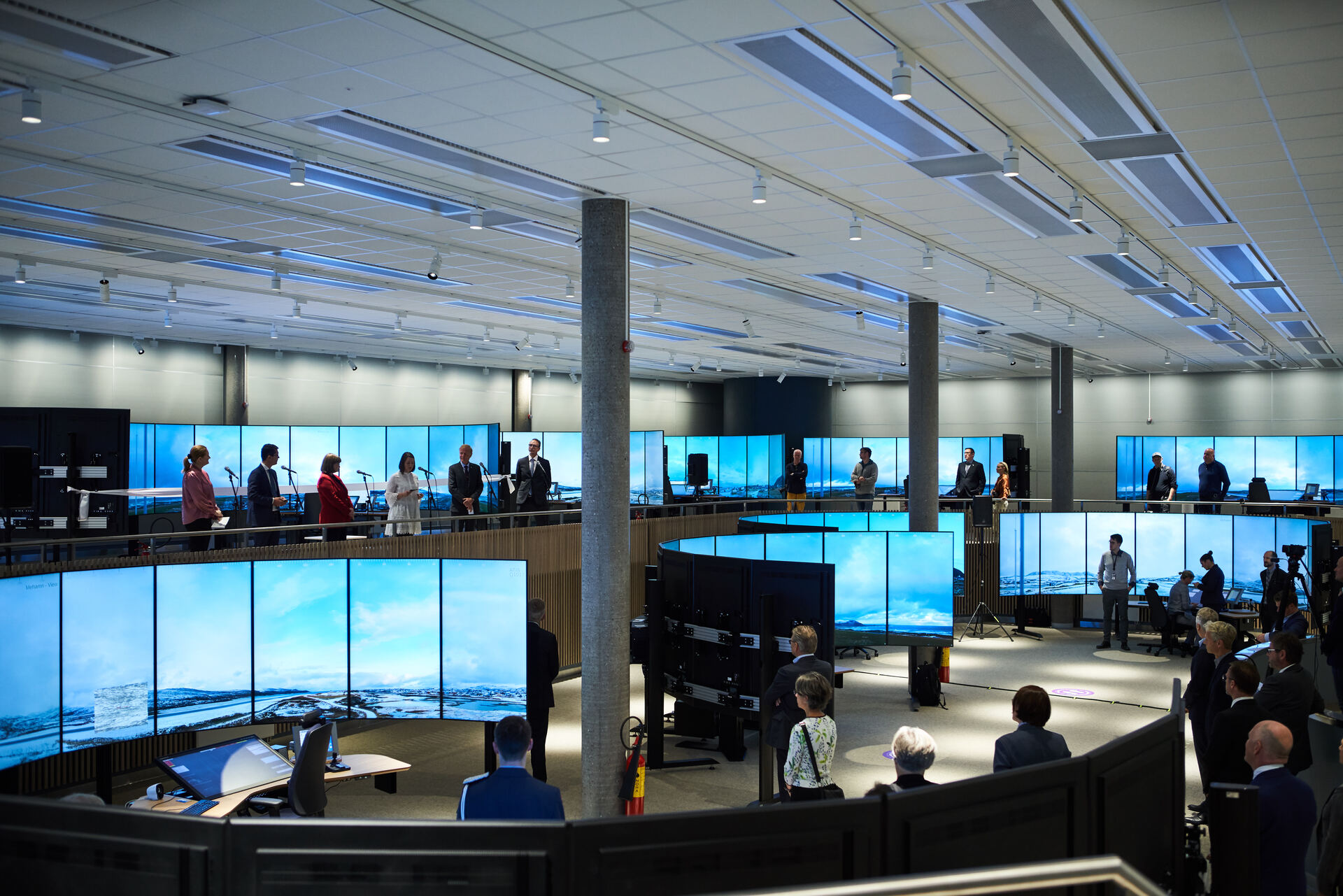 Spacious modern control room with multiple large display screens and staff members standing, showcasing operational monitoring and management.