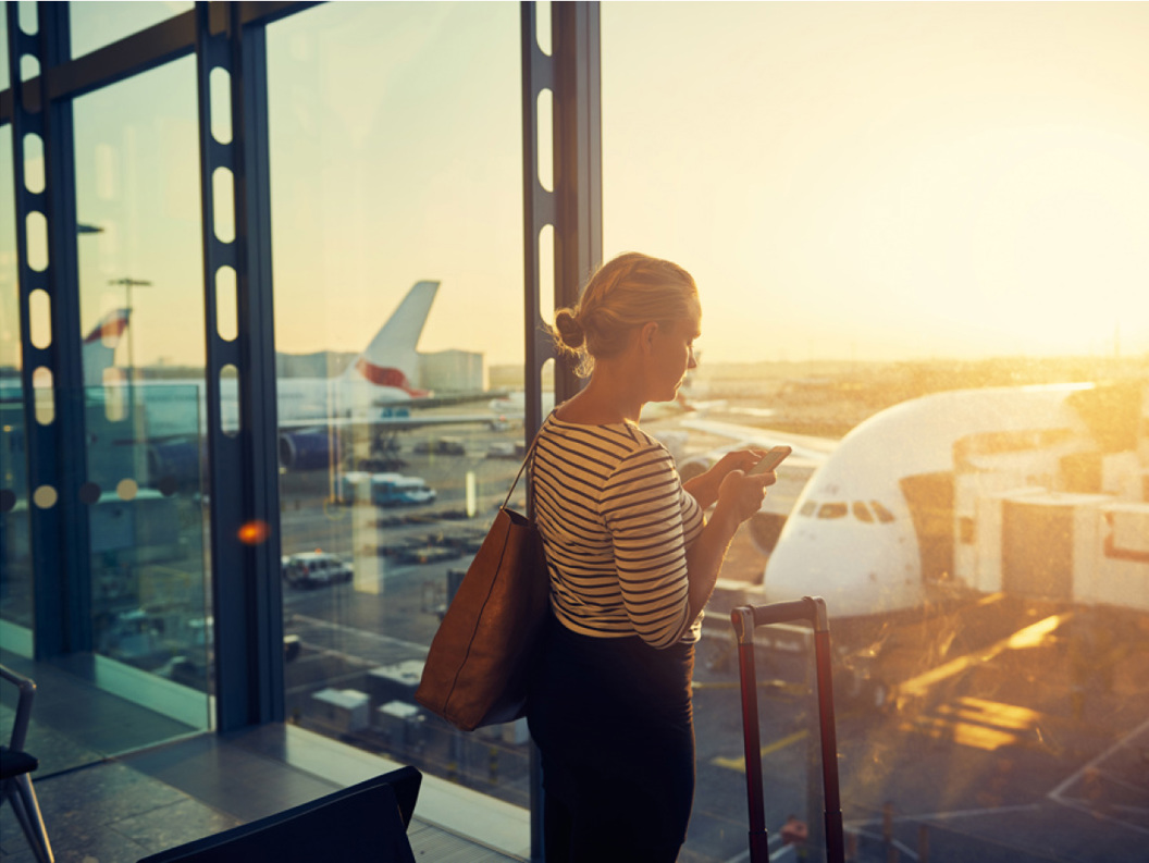 Reisende bruker smarttelefon på flyplassterminal med fly i bakgrunnen under solnedgang