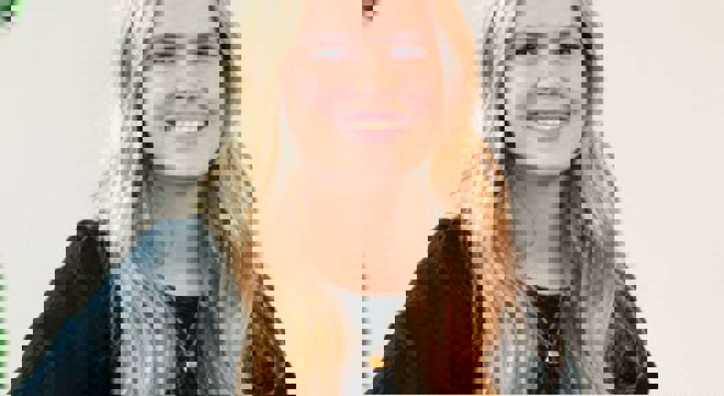 Smilende kvinne med langt blondt hår som har på seg en svart topp, sittende innendørs med en plante i bakgrunnen