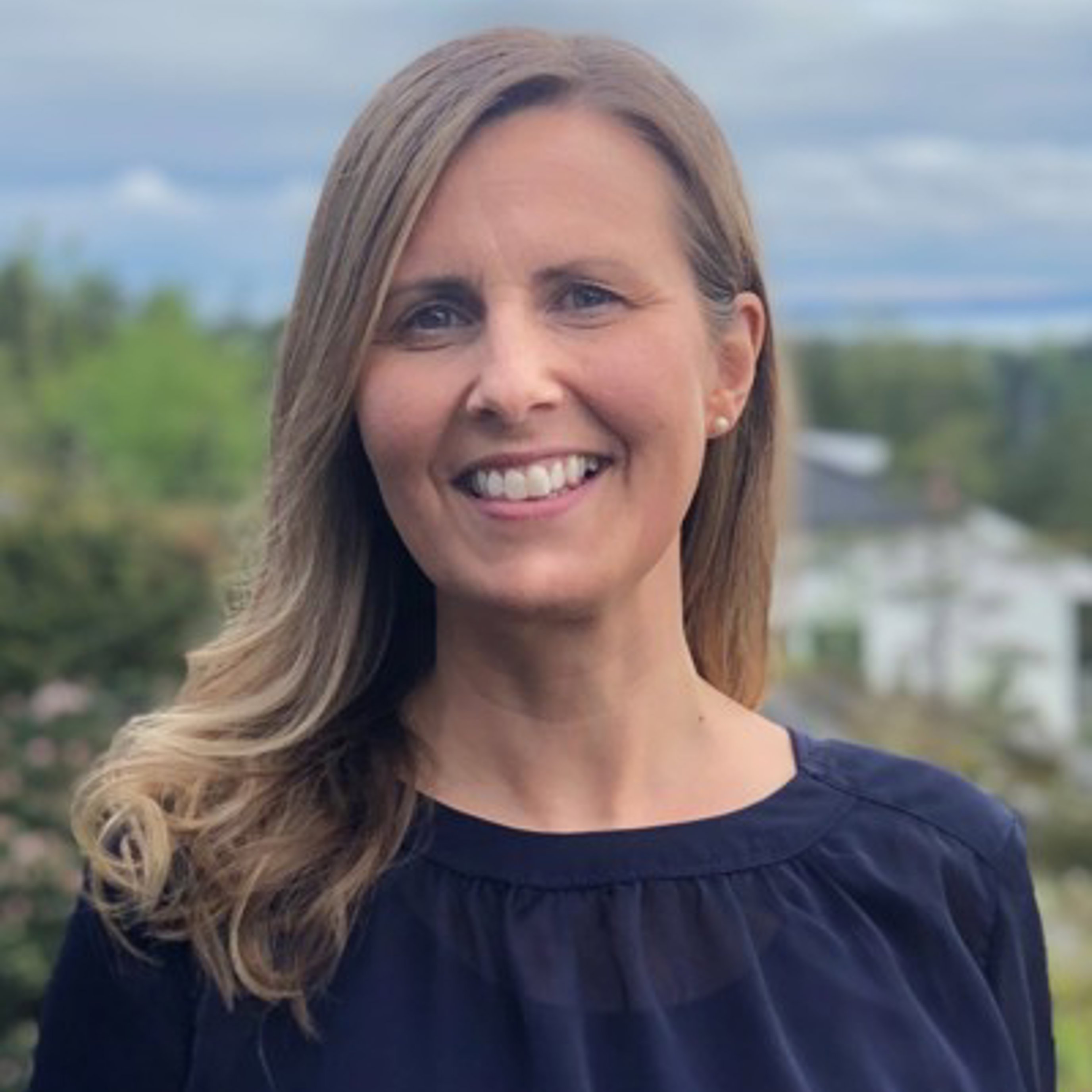 Kvinne med langt hår som smiler, iført en marineblå bluse, stående utendørs med en uskarp naturlig bakgrunn