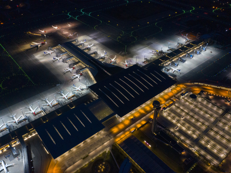 Flyfoto av opplyst flyplass med parkerte fly og terminalbygning om natten.