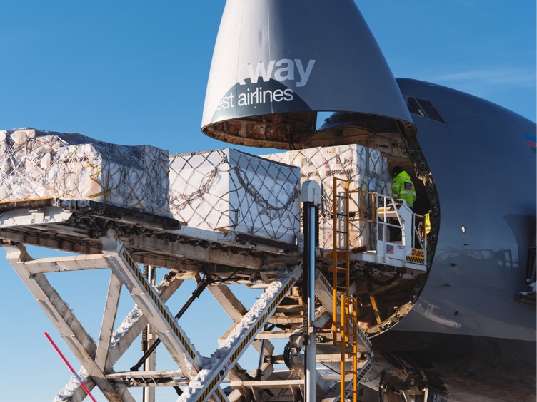 Aircraft undergoing cargo-handling work at a snow-covered airport. Raised access platform, exposed nose section, scaffolding and winter mountain landscape surround aircraft beneath clear blue sky.