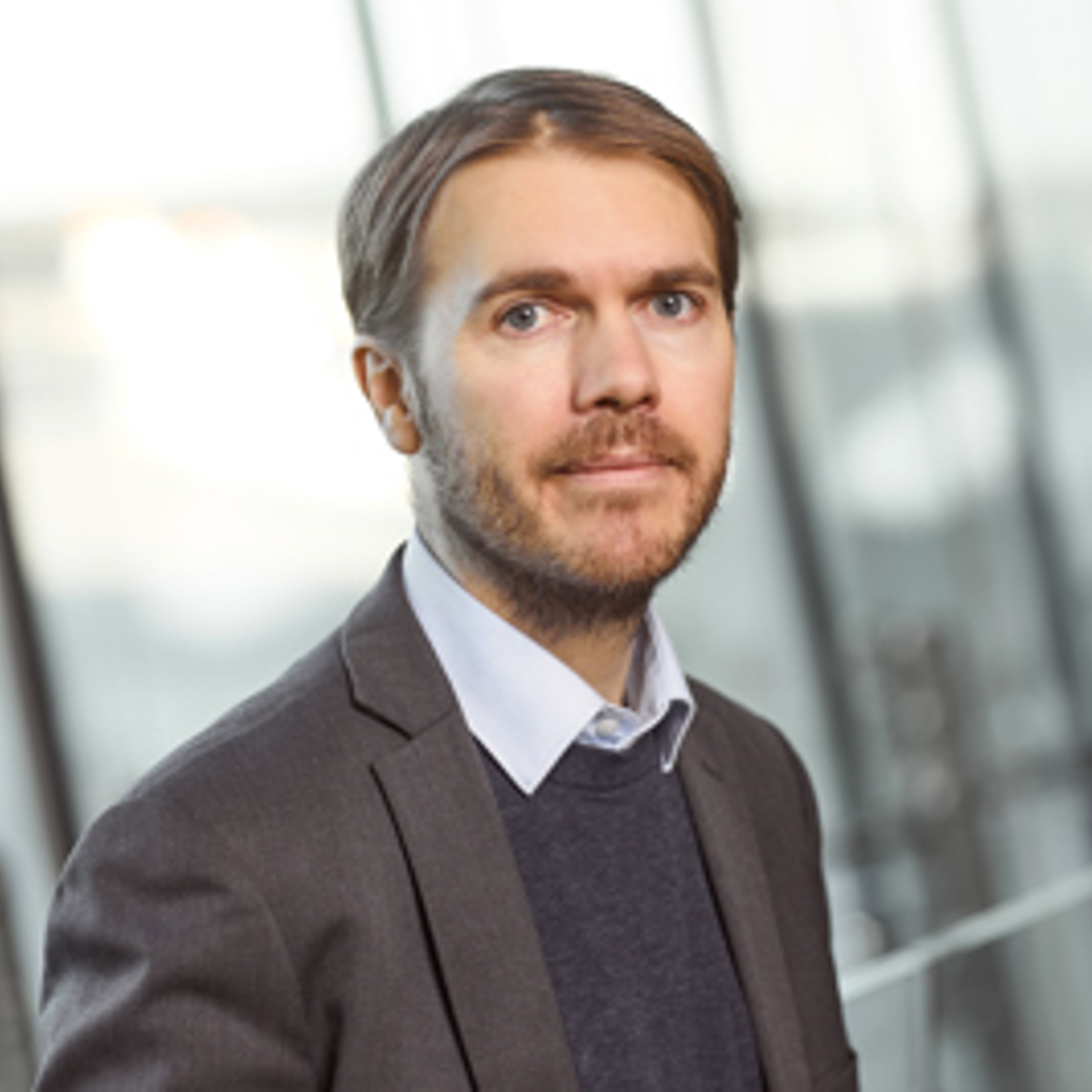 Mann i dress med et alvorlig uttrykk som står innendørs nær store vinduer