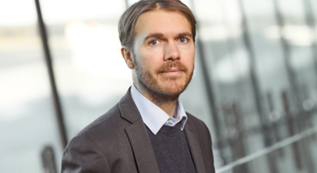 Mann i dress med et alvorlig uttrykk som står innendørs nær store vinduer