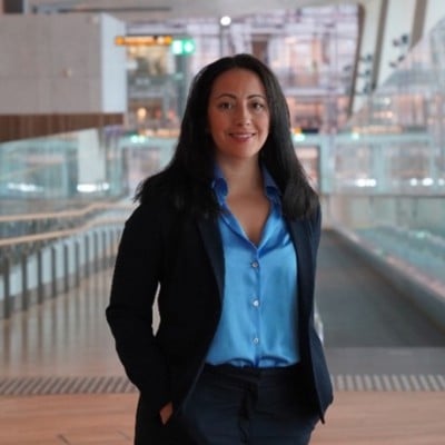 Woman in blue shirt and dark blazer stands in a modern building with glass walls.
