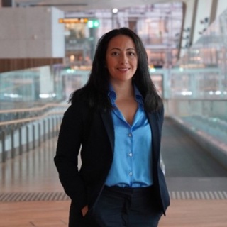 Woman in blue shirt and dark blazer stands in a modern building with glass walls.