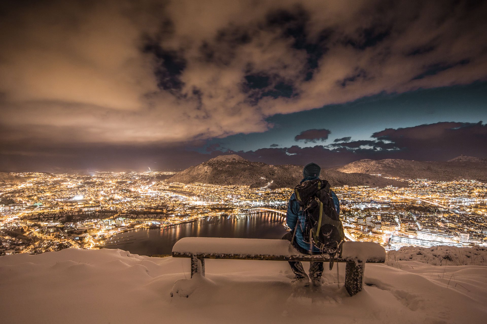 Person på snødekket benk med utsikt over opplyst Bergen by om natten, med fjell i bakgrunnen.