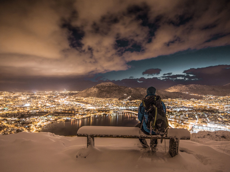 Person på snødekket benk med utsikt over opplyst Bergen by om natten, med fjell i bakgrunnen.