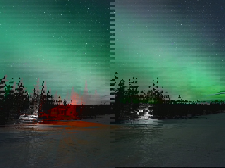 Nordlys over snødekt landskap med en isolert hytte omgitt av skog i vinterkveld.