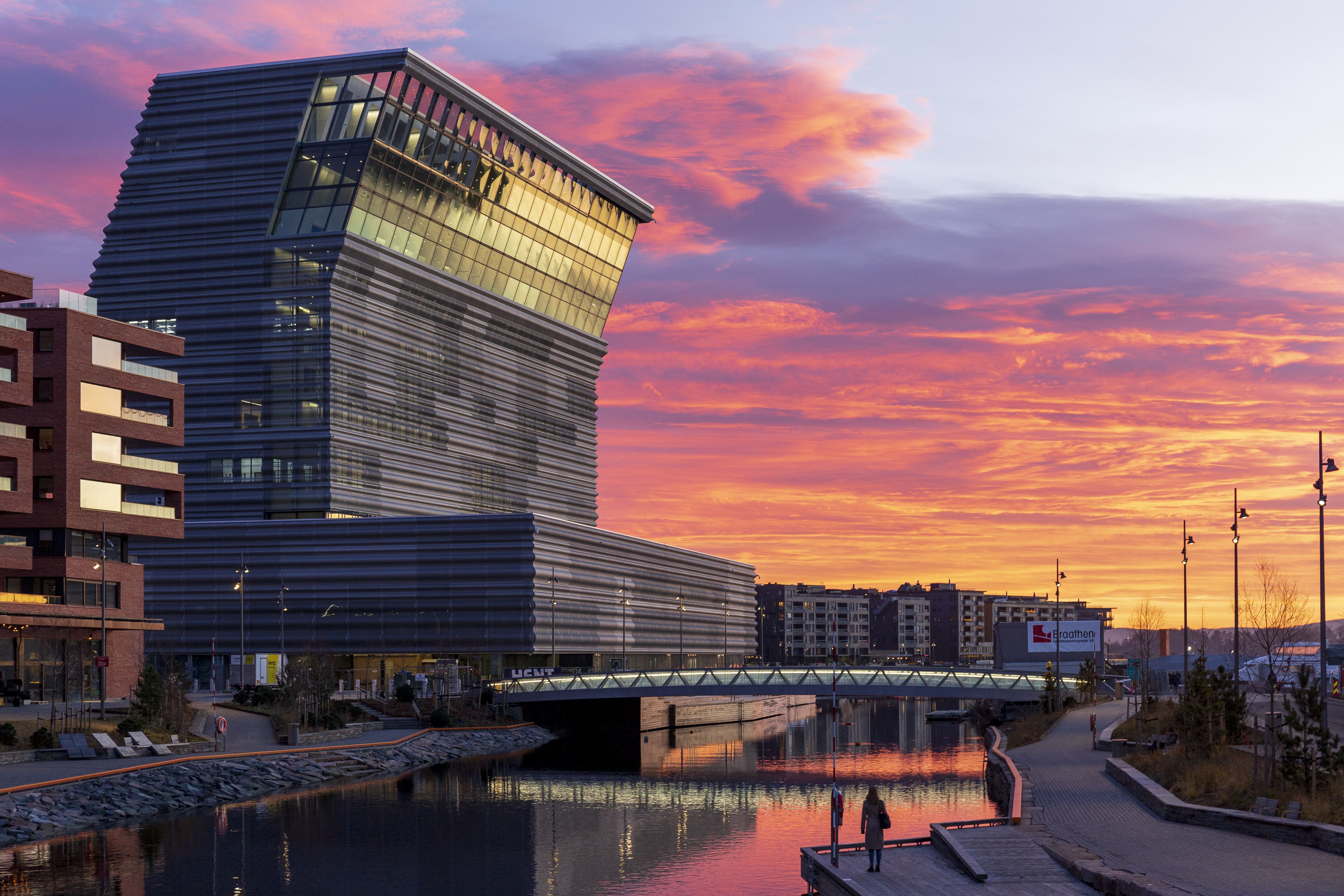 Munchmuseet i Oslo mot en dramatisk solnedgang, med moderne arkitektur speilet på vannoverflaten.