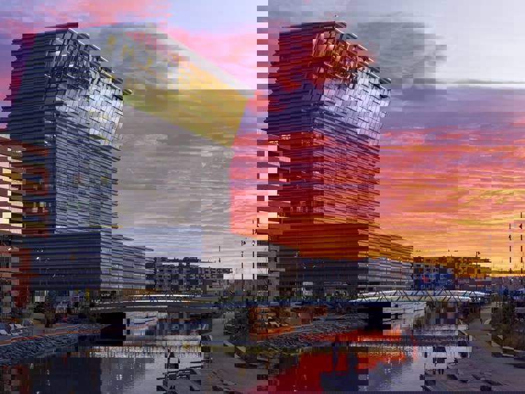 Munchmuseet i Oslo mot en dramatisk solnedgang, med moderne arkitektur speilet på vannoverflaten.