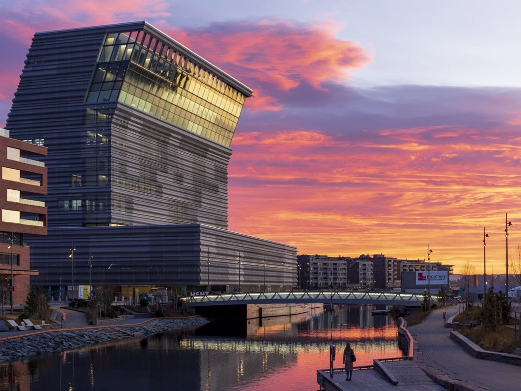 Munchmuseet i Oslo ved solnedgang, med dramatiske rosa og oransje skyer i bakgrunnen, reflektert i vannet nedenfor.