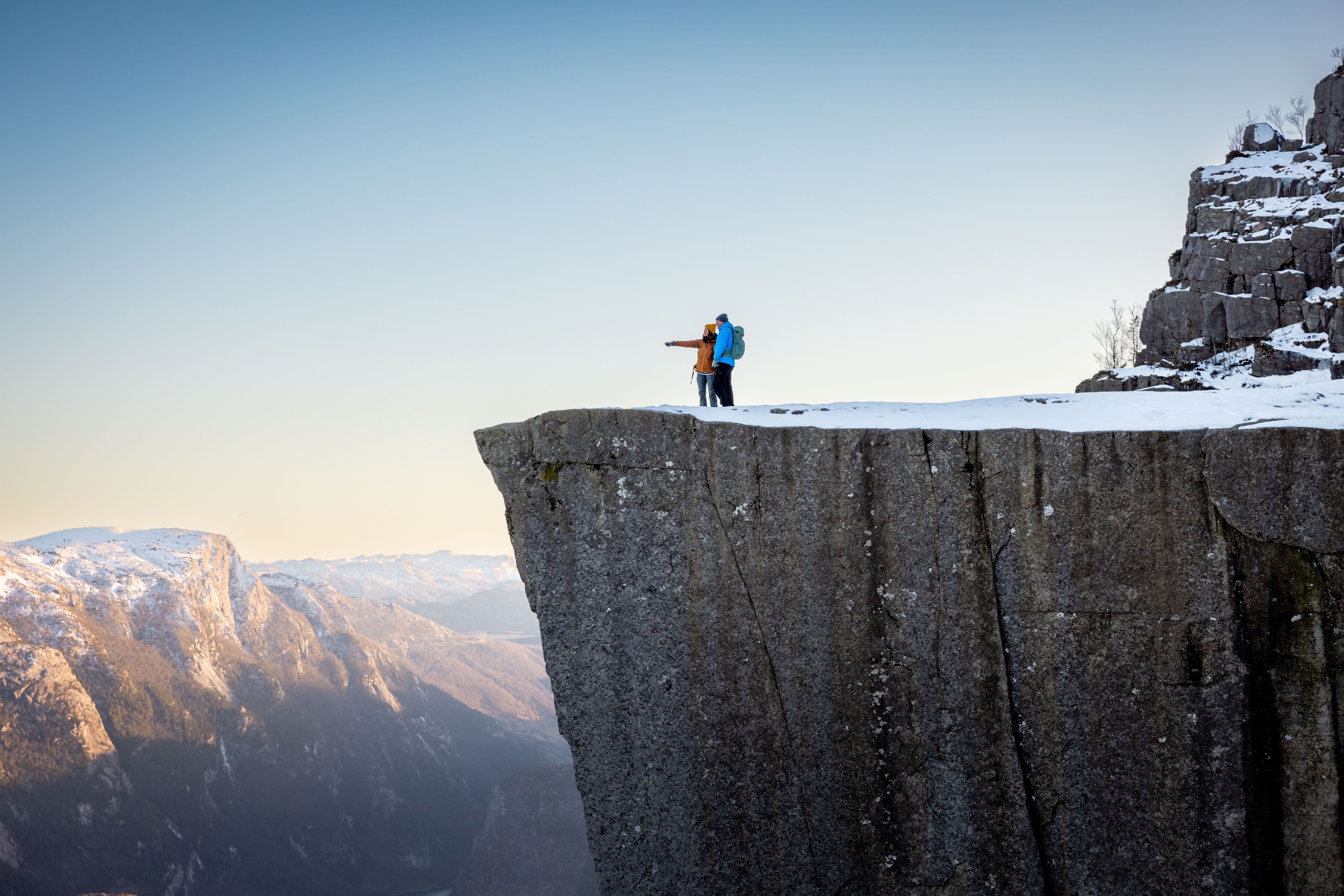 To personer står på kanten av Preikestolen, en kjent klippe i Norge, med utsikt over snødekte fjell og en klar himmel.