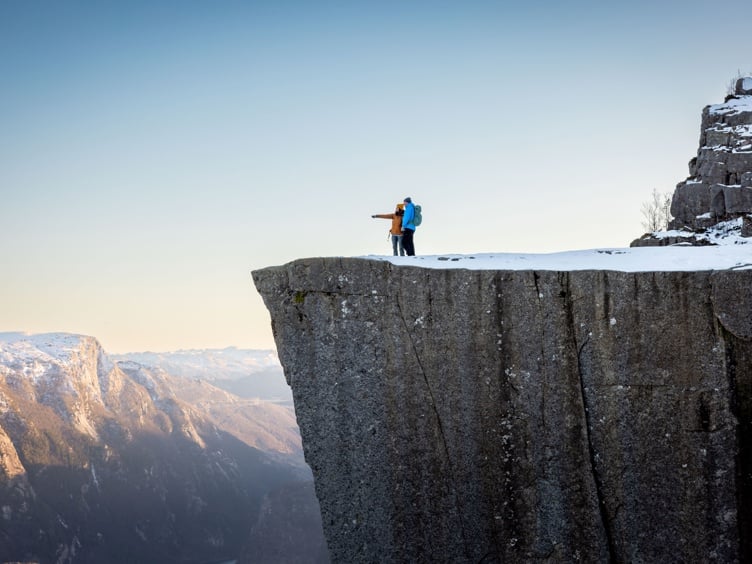 To personer står på kanten av Preikestolen, en kjent klippe i Norge, med utsikt over snødekte fjell og en klar himmel.