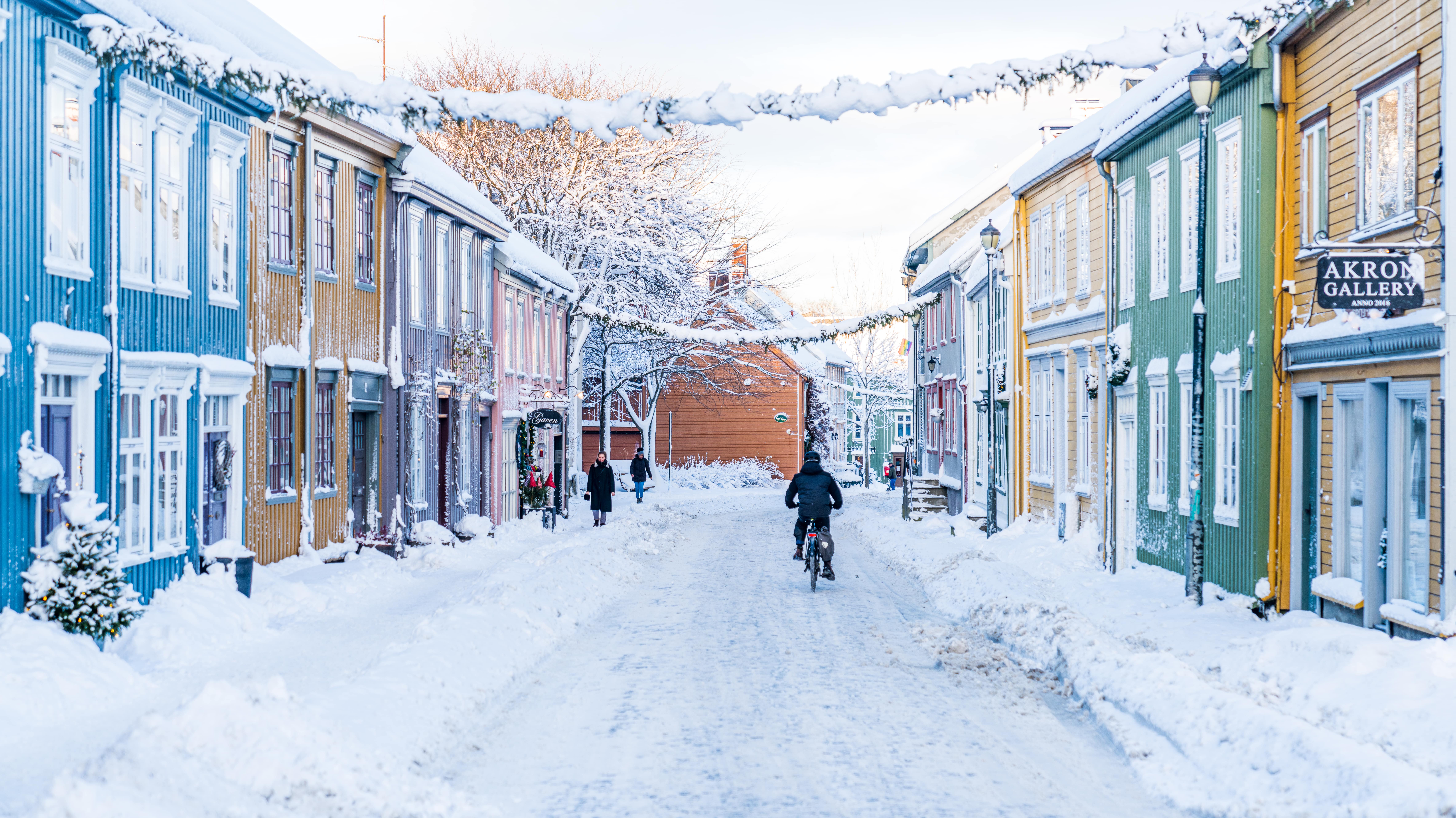 Snødekt gate i et livlig nabolag med fargerike trehus, en person sykler langs veien og andre går forbi i vinterlandskapet.