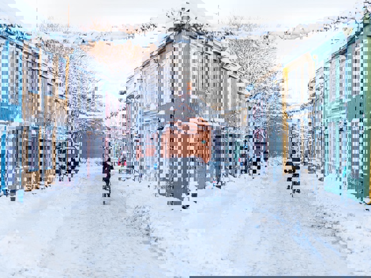 Snødekt gate i et livlig nabolag med fargerike trehus, en person sykler langs veien og andre går forbi i vinterlandskapet.